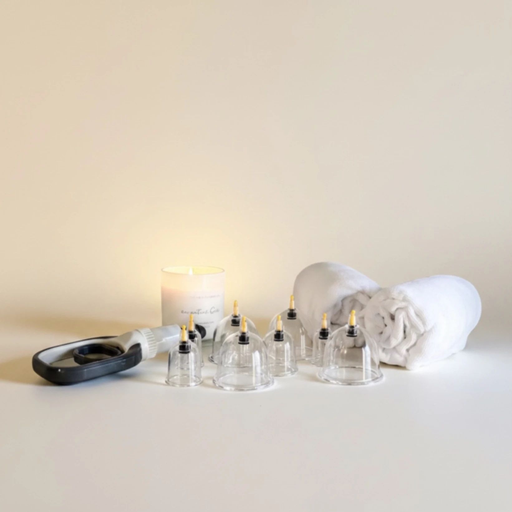 Cupping cups, a pump, a lit candle and rolled white towels on a cream surface