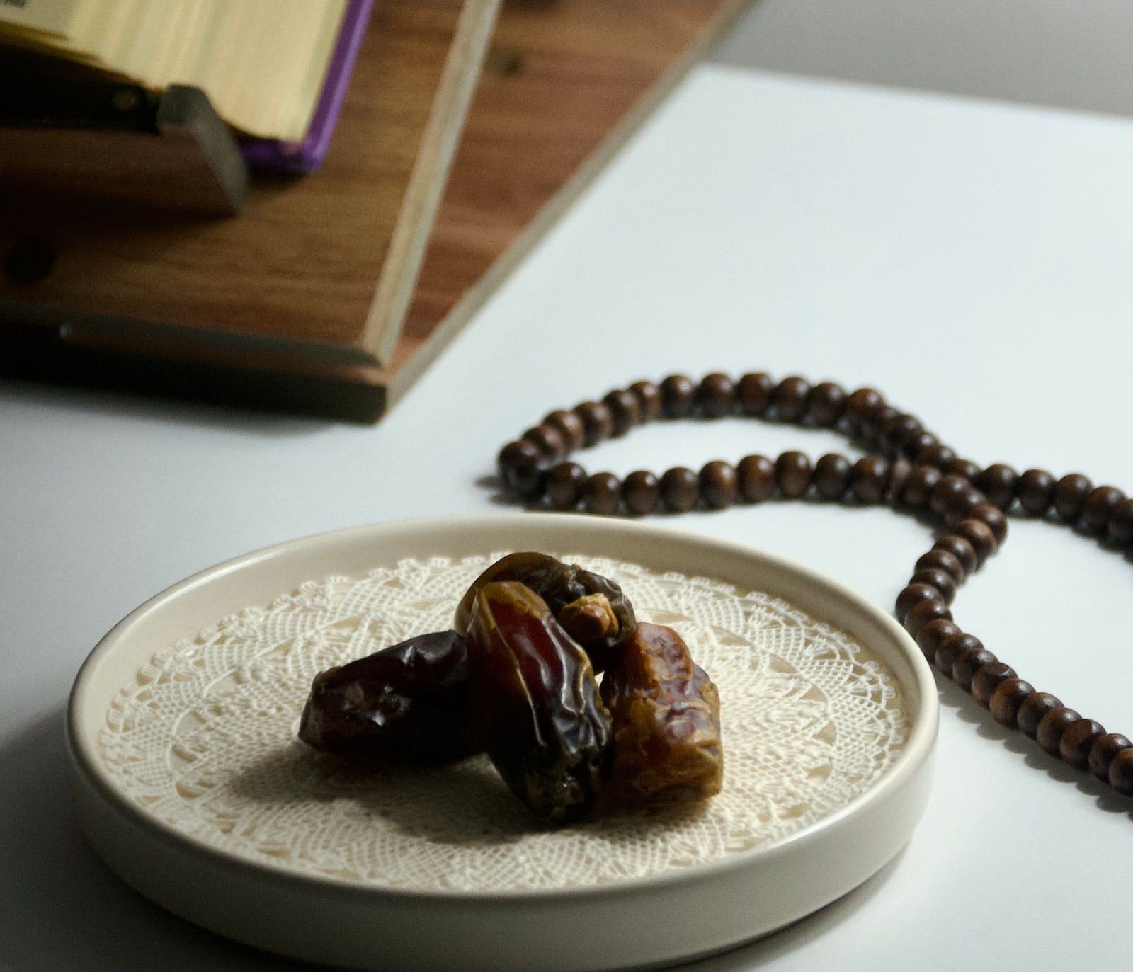 A Quran resting on a wooden stand, with wooden prayer beads and dates on a lace-lined plate