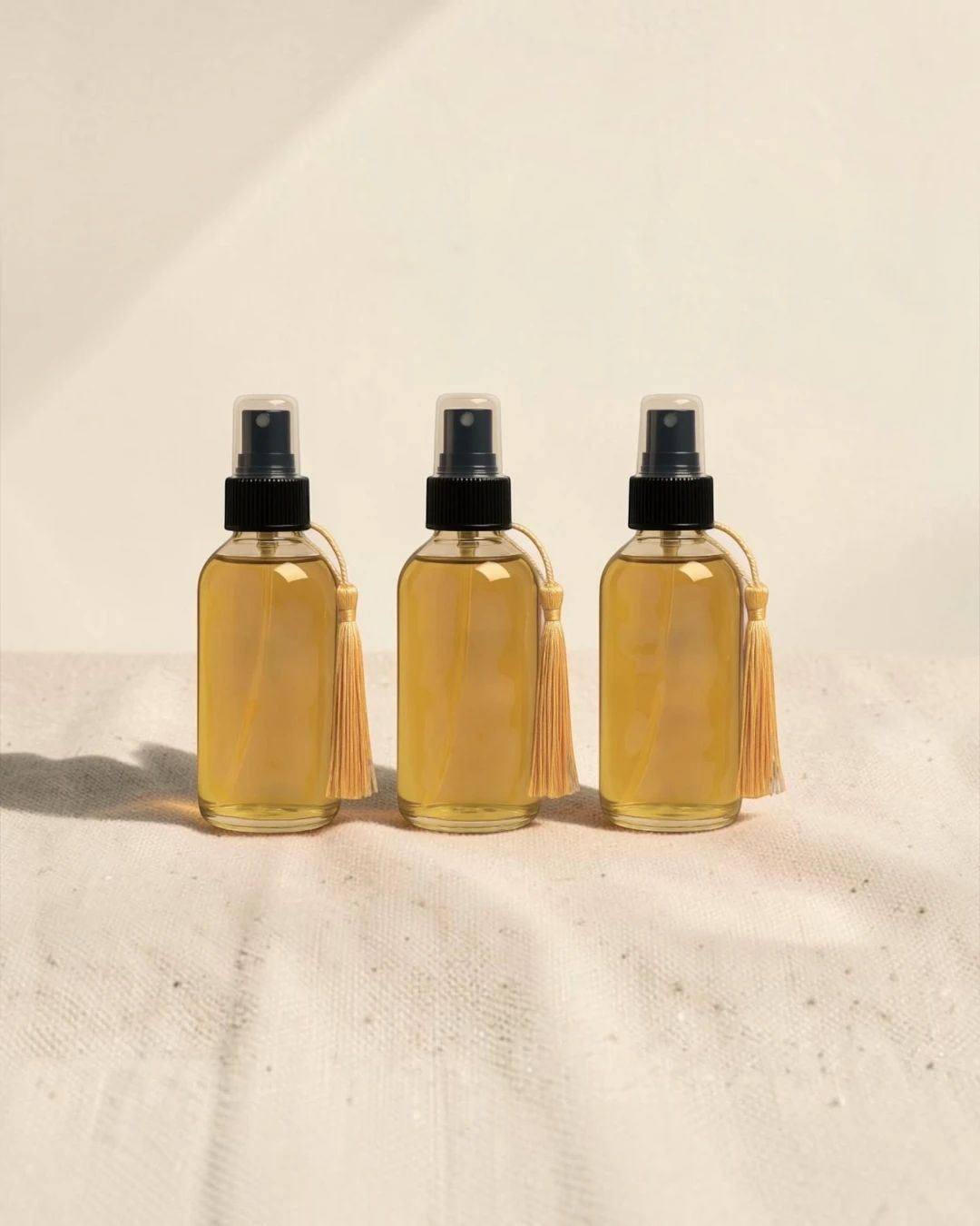 Three glass spray bottles of Ruqyah oil with gold tassels on cream linen